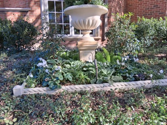 A curved stone border outlines mixed foliage and flowers around a garden urn.