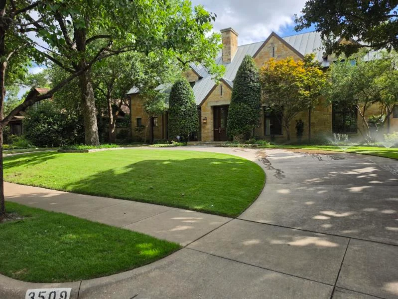 Operating sprinklers water a lawn bordered by mature trees and foundation planting.