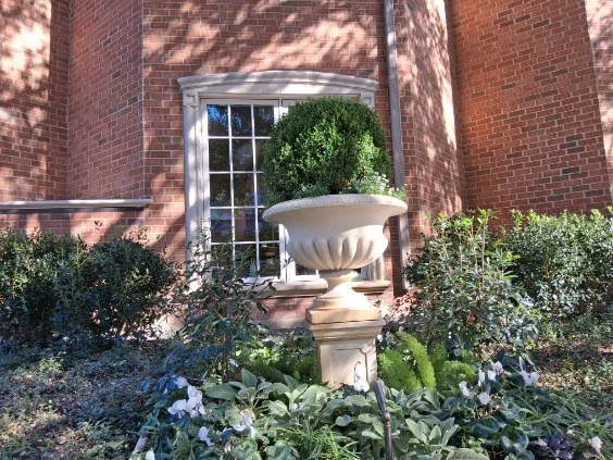 A planted stone urn stands among mixed foliage beneath a window.