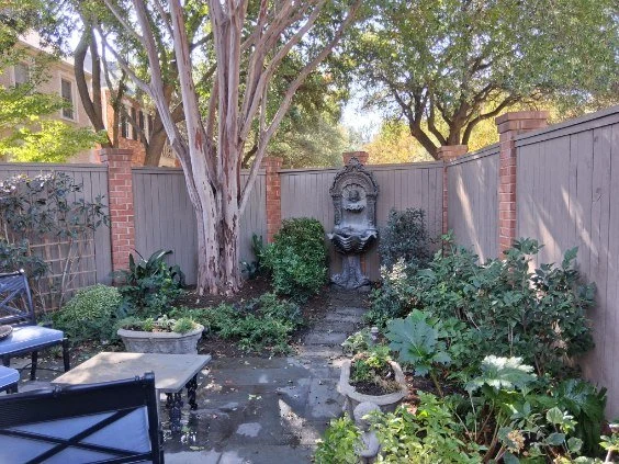 A mature tree, mixed planting, and a decorative fountain surround a paved seating area.