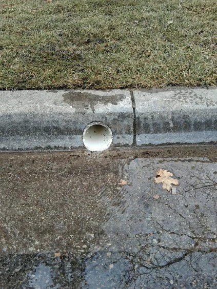 A round drainage outlet discharges through the curb at the edge of a lawn.