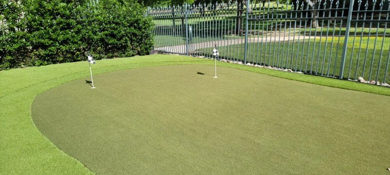 A compact putting surface is framed by turf, planting, and a metal fence.
