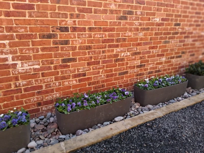 Rectangular flower planters sit among smooth stones along a brick wall.